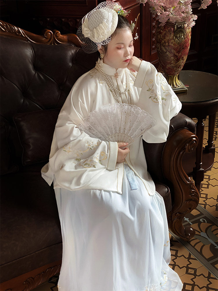 Woman in traditional white dress with lace details, holding a fan, sitting on a leather chair.